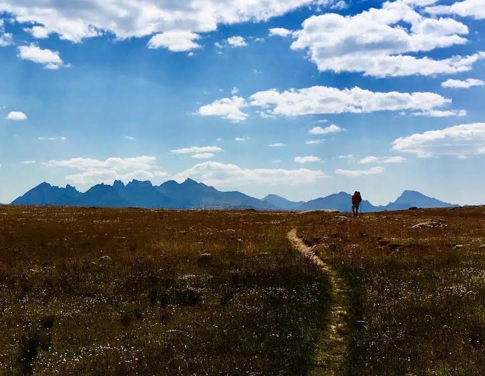 A backpacker stands on the horizon, in front of a single track trail through a meadow. Peaks loom in the distance.