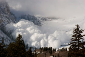 A plume of snow from an avalanche rises into the sky on a snowy peak.
