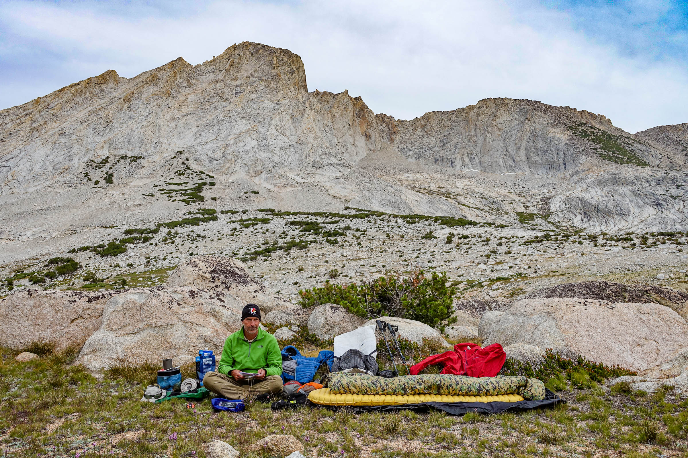 Adventure Alan sits with all of his backpacking gear spread out around him in front of some barren peaks.