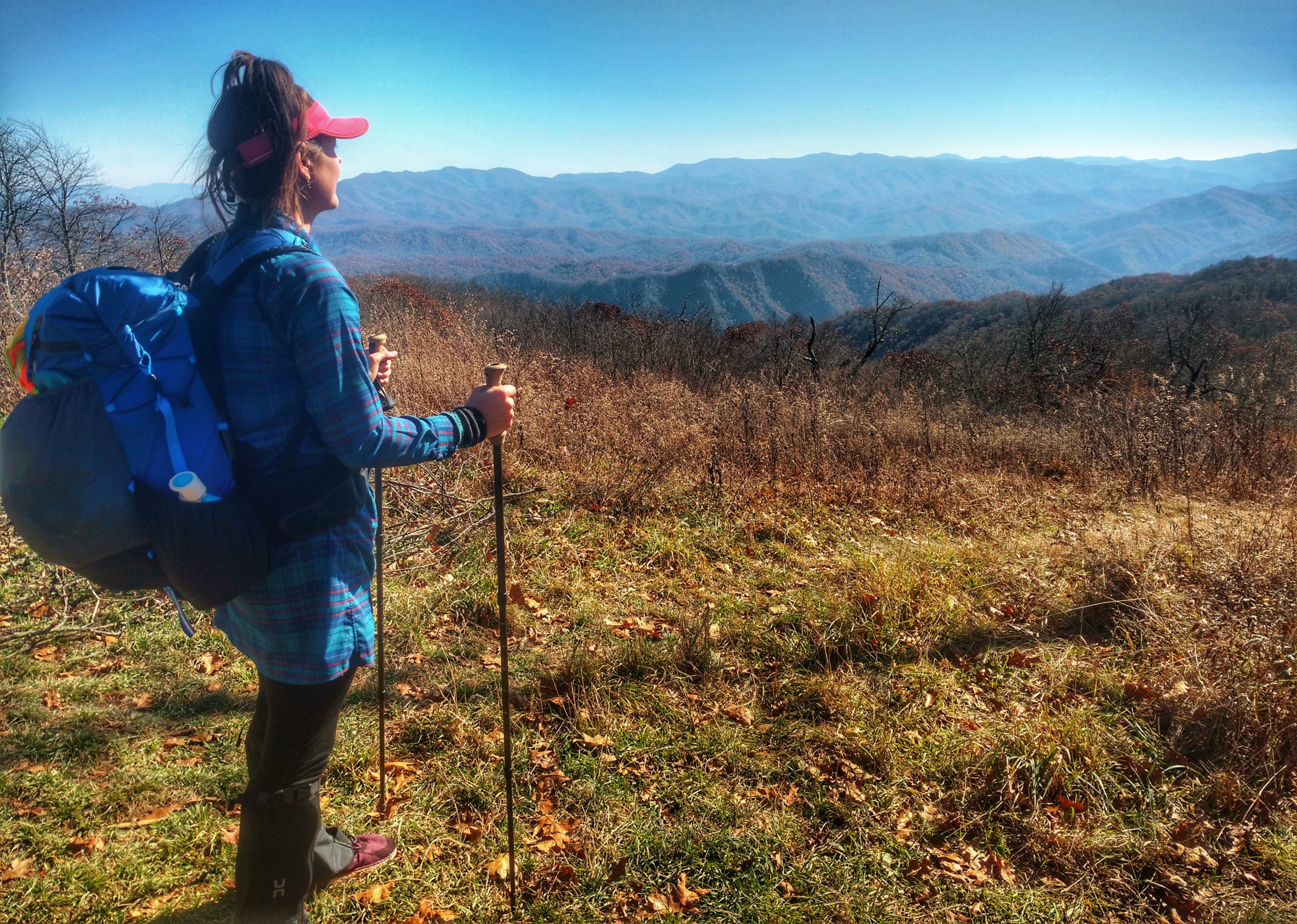 Anish stands with her poles and backpack, gazing out from a field to a rainbow sea of mountains.
