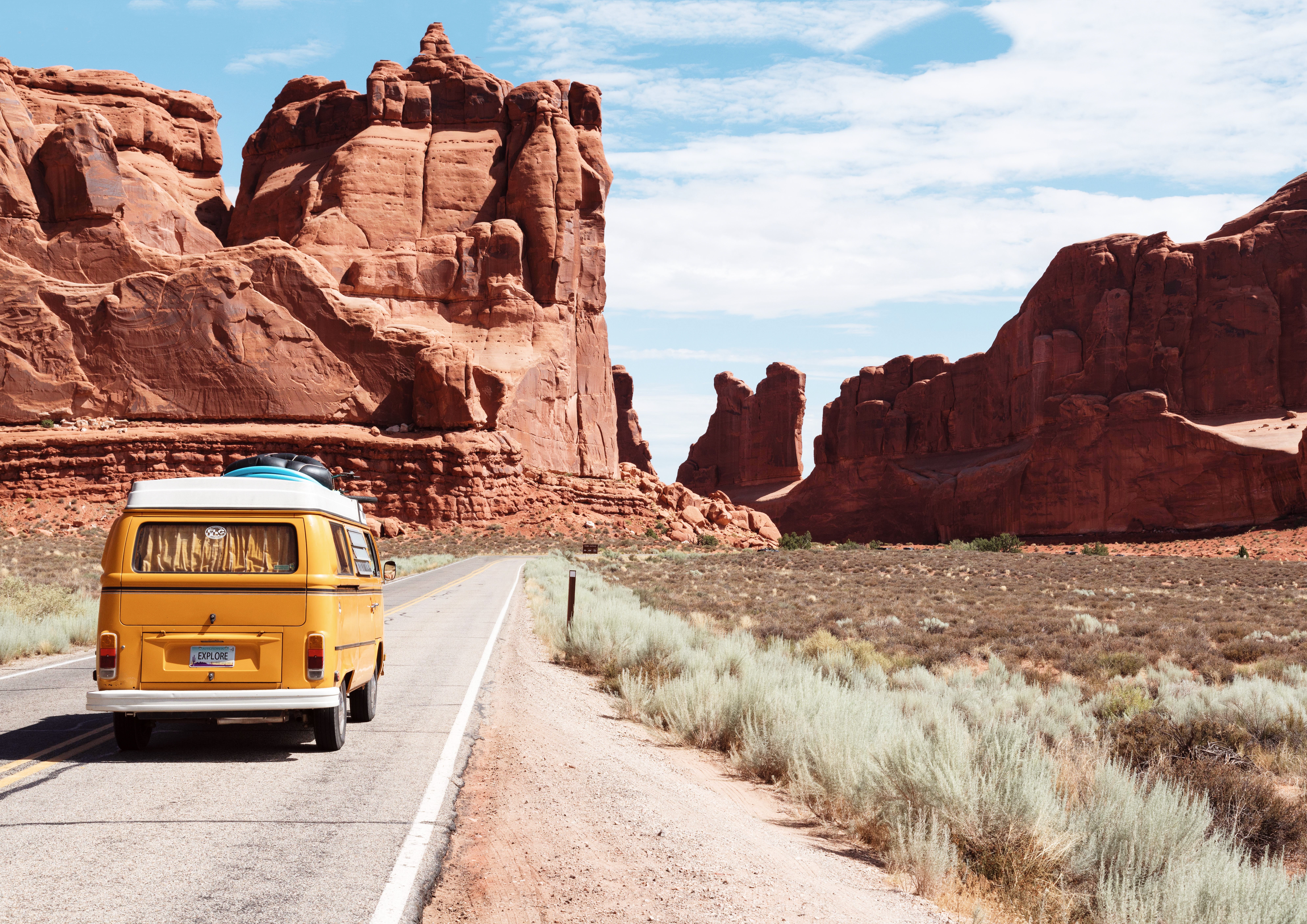 A yellow camper bus drives down a desert road towards red-walled rock towers. 