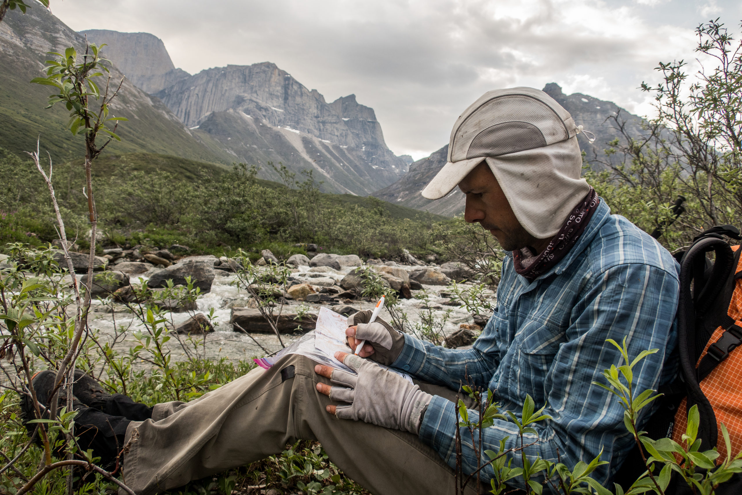 Skurka sits with his back against a backpack while taking notes. A river flows behind him, and peaks loom in the distance.