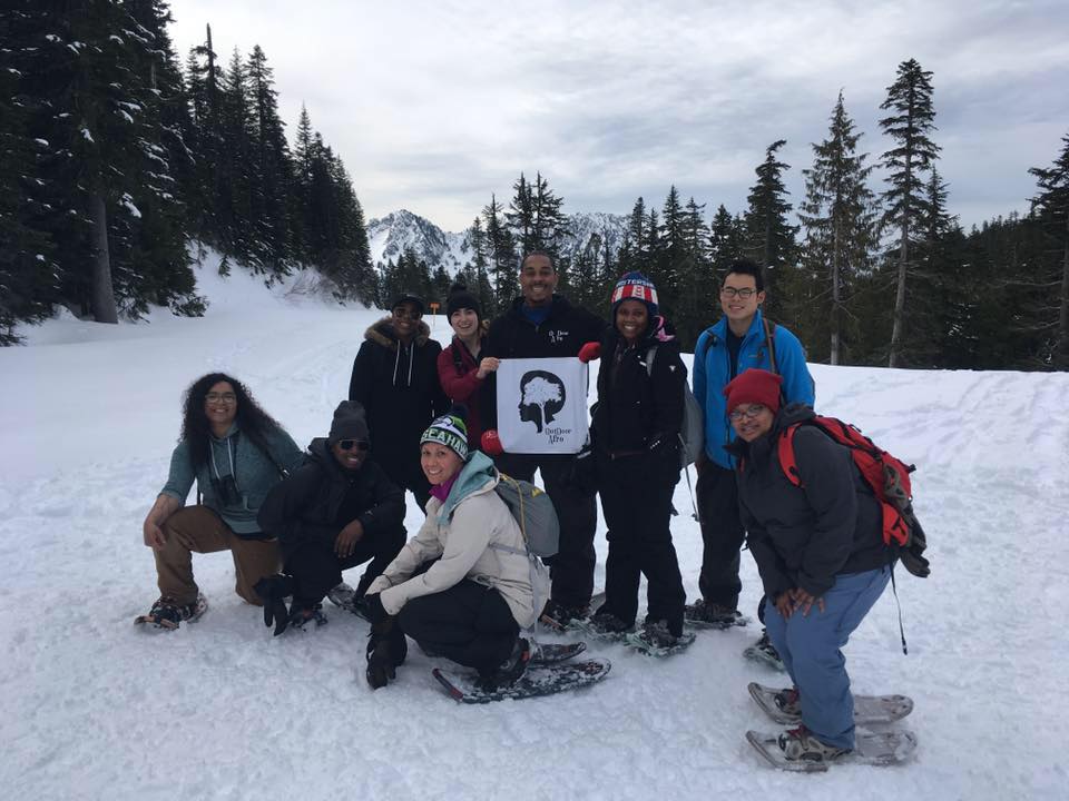 A group of snowshoers smile for the camera on a snow-covered trail. Someone in the middle is holding a sign with the Outdoor Afro logo. 