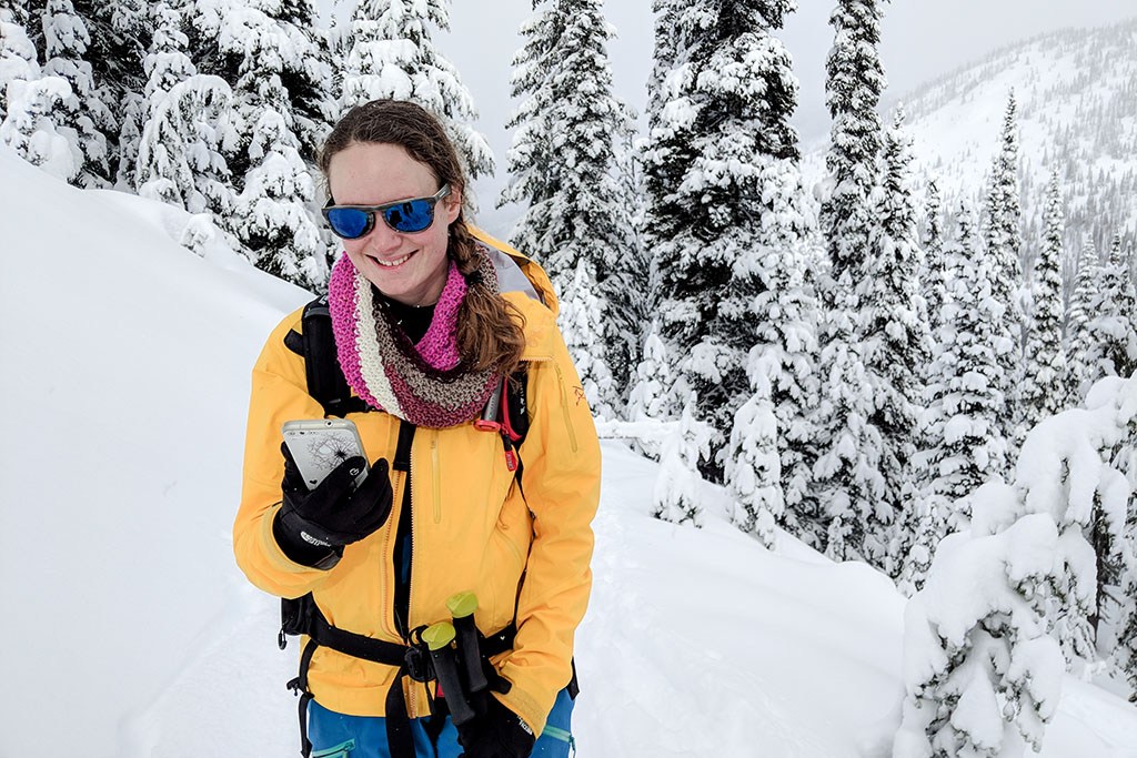 A backcountry skier smiles while looking at her phone. She stands in front of a row of snow-covered coniferous trees.