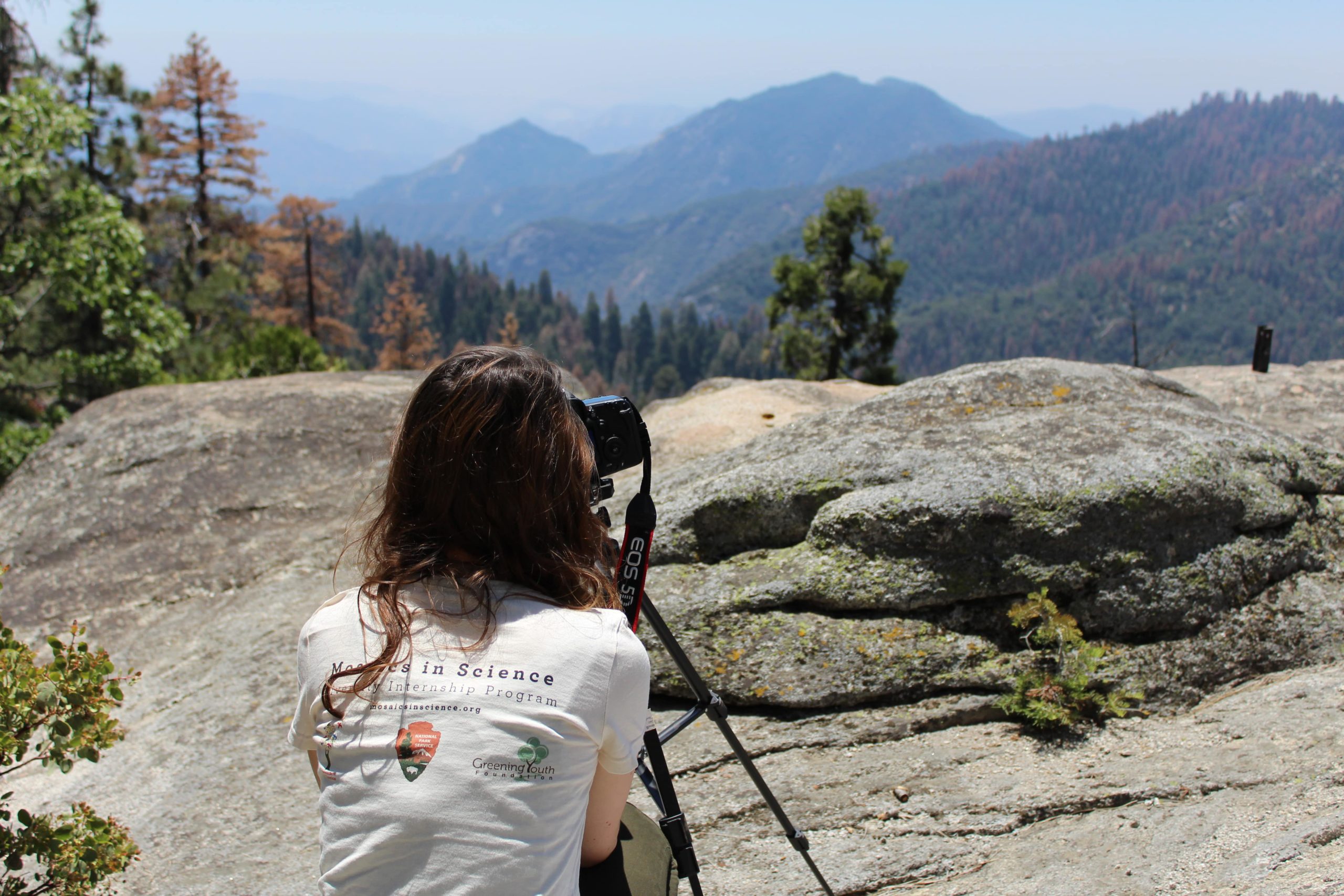 A GYF intern looks through a camera set up on a tripod. She's sitting on a rocky outcropping, gazing at mountains before her.