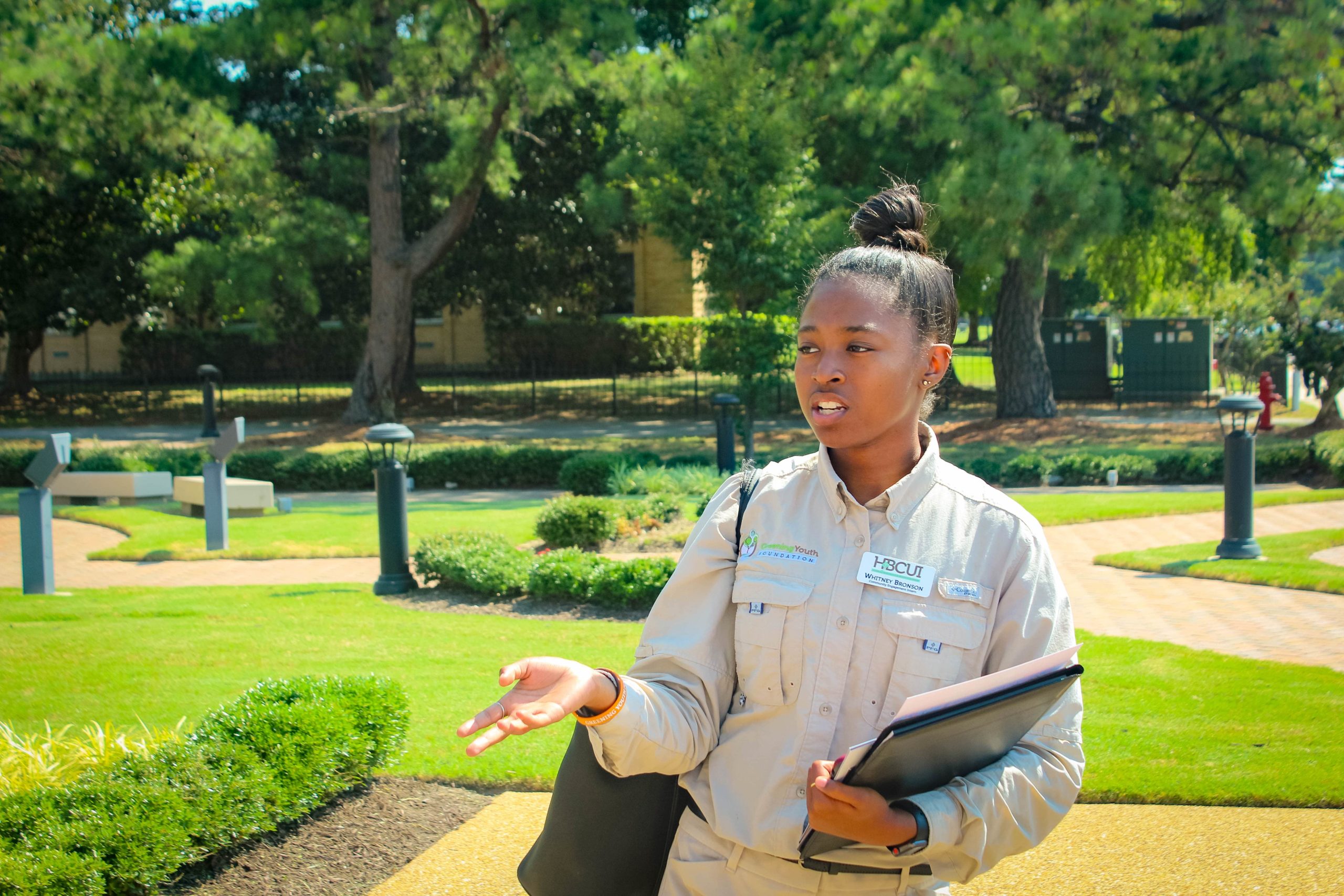 A GYF intern holds a clipboard in one hand and extends her other hand out. She stands in a sunny, manicured grassy park.