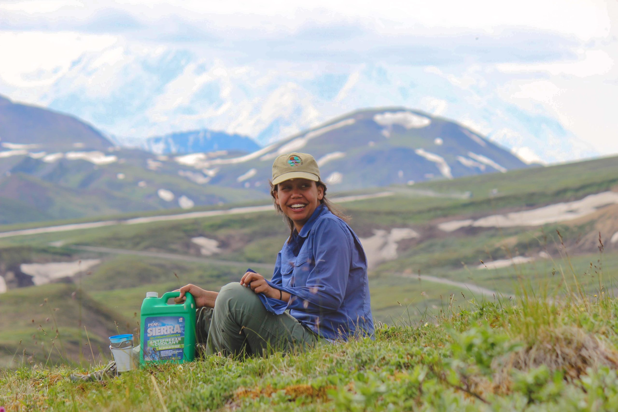 A GYF intern smiles while sitting in a field and holding a bottle of anti-freeze. Snowcapped peaks rise into a cloudy sky behind her.
