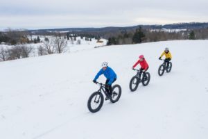 Three people in brightly colored jackets ride fat bikes on a snowy trail.