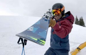 A skier holds up his ski to the camera. He stands on a ski slope, and is wearing a helmet and googles.
