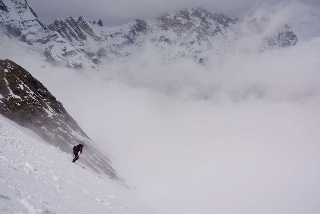 Smithwick skis down a steep wind-swept slope. Tall jagged peaks are visible above clouds of snow in the background.