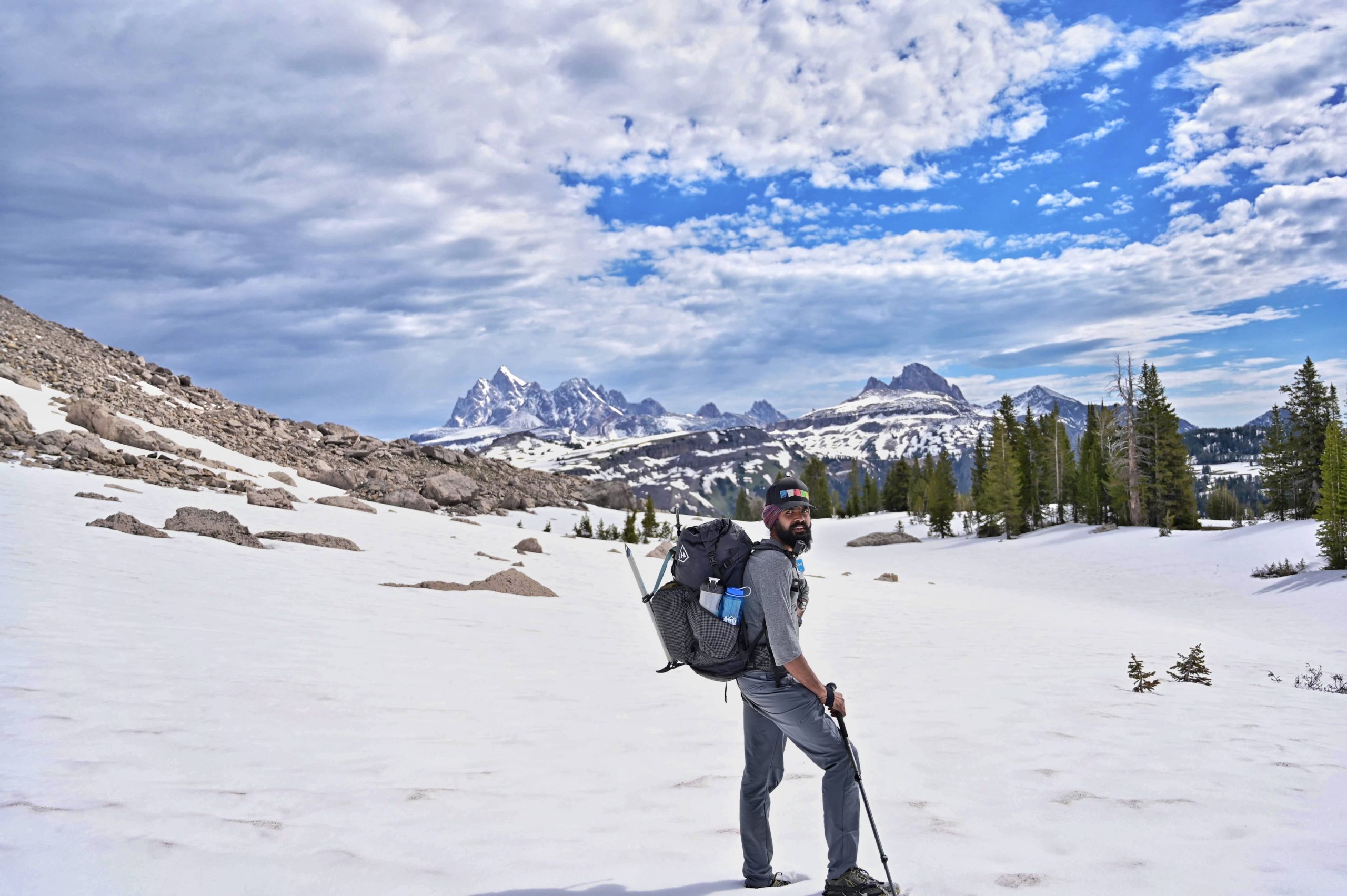 Anu stands in a snowfield with jagged peaks in the background. He's holding hiking poles and carrying a big backpack.