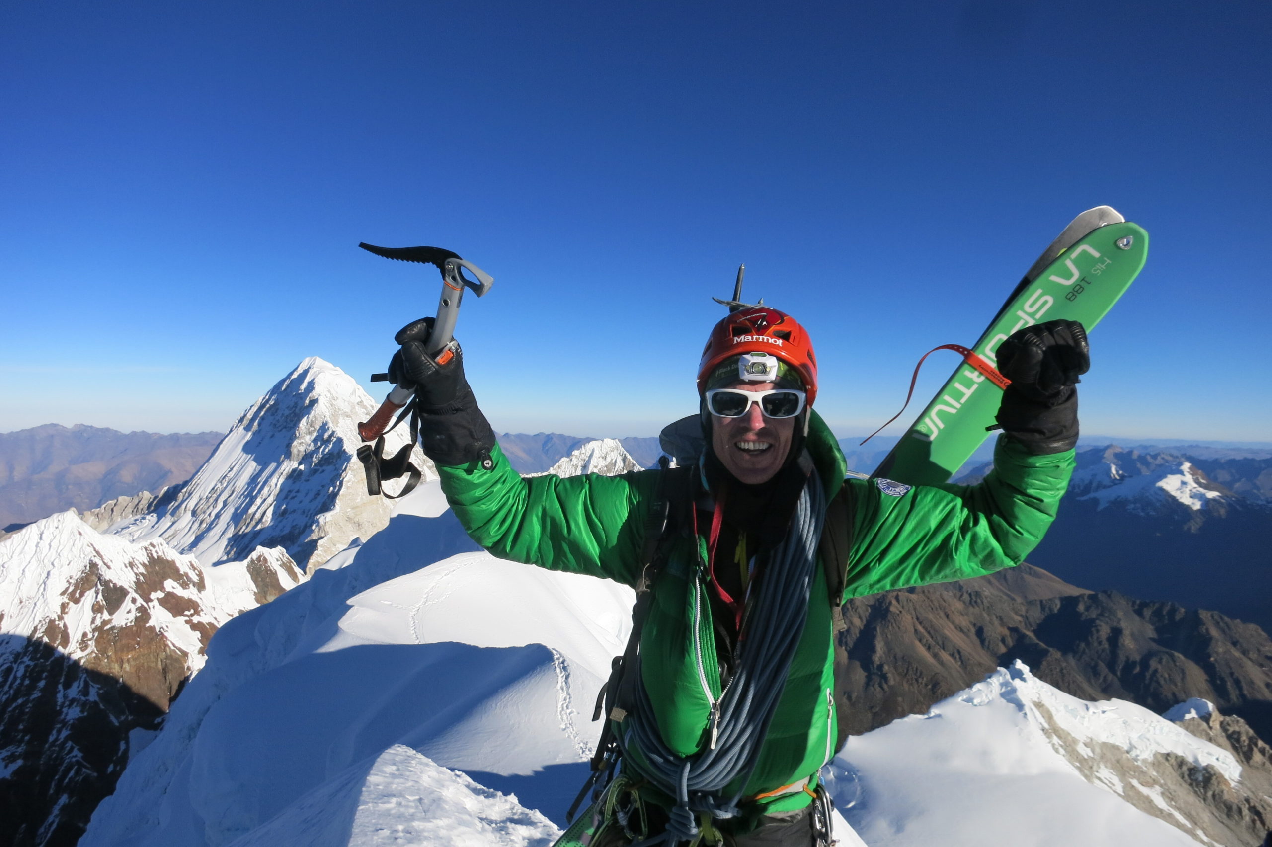 Adrian stands on top of a mountain holding an ice axe in one hand and his gloved fist raised in the other. His skis are on his back. He's wearing sunglasses, a headlamp, and a helmet. Snowcapped speaks are all around him in the background.