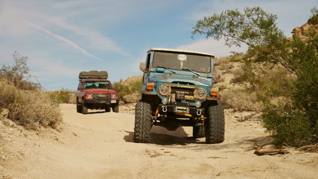 Old blue offroad vehicle followed by a read vehicle drive through the sand. 
