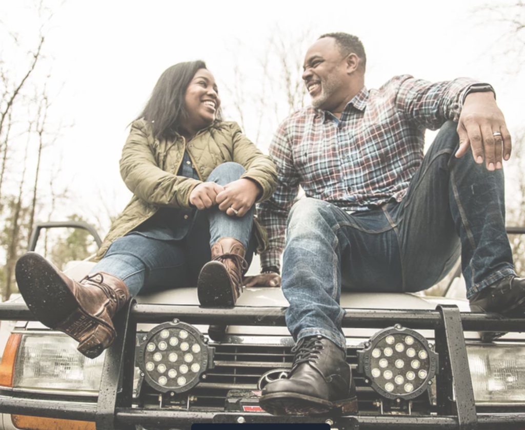 A man and a woman are sitting on the hood of their truck, smiling and looking at each other with love.