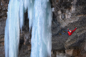 An ice climber hangs from an overhanging rock wall with a frozen waterfall beside him.