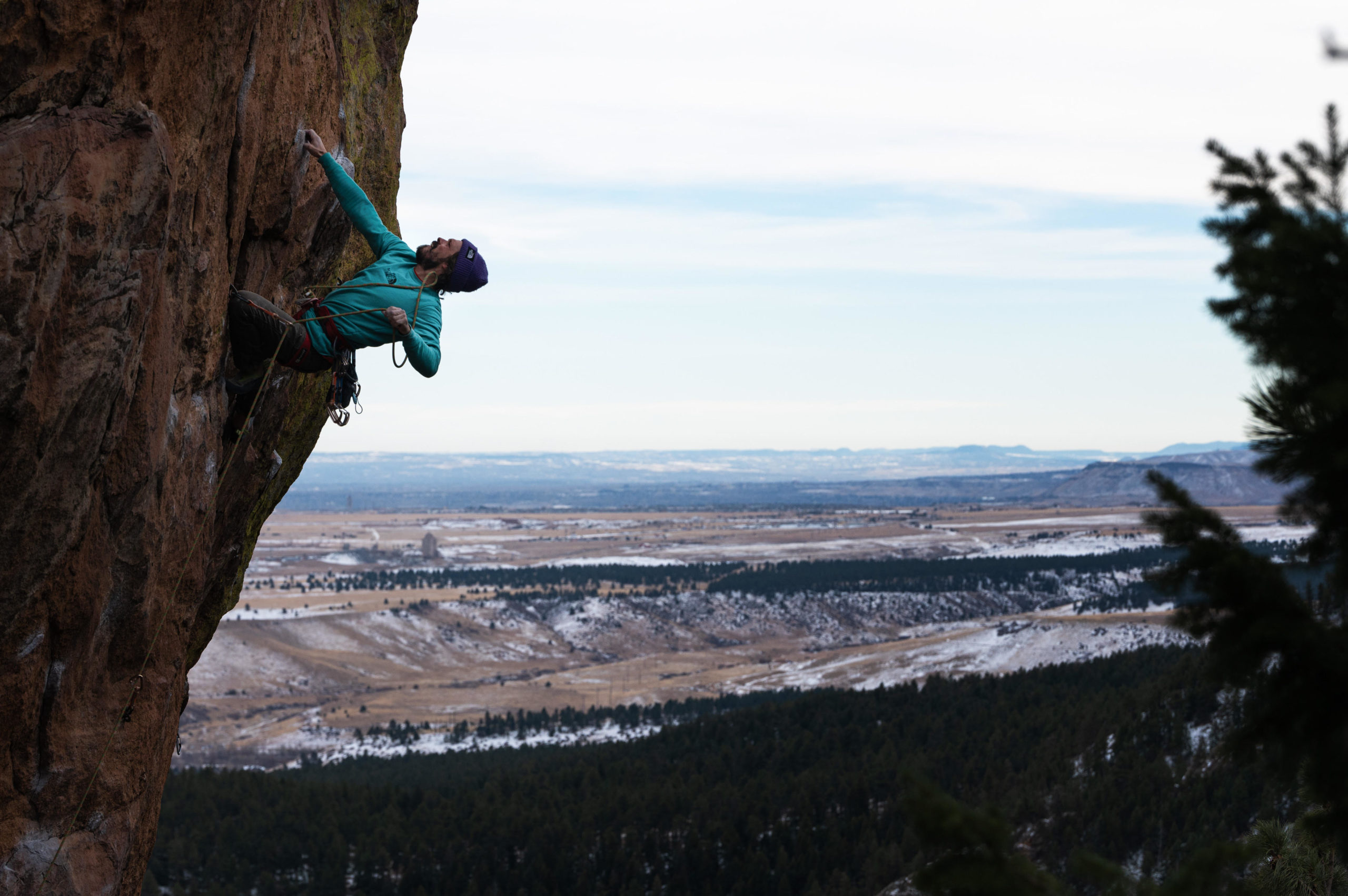 Segal is hanging onto a rock wall with one hand. His feet are placed on the rock below, and his other hand is pulling the rope attaching him to the wall up to clip it in higher. Snowy plains extend behind.
