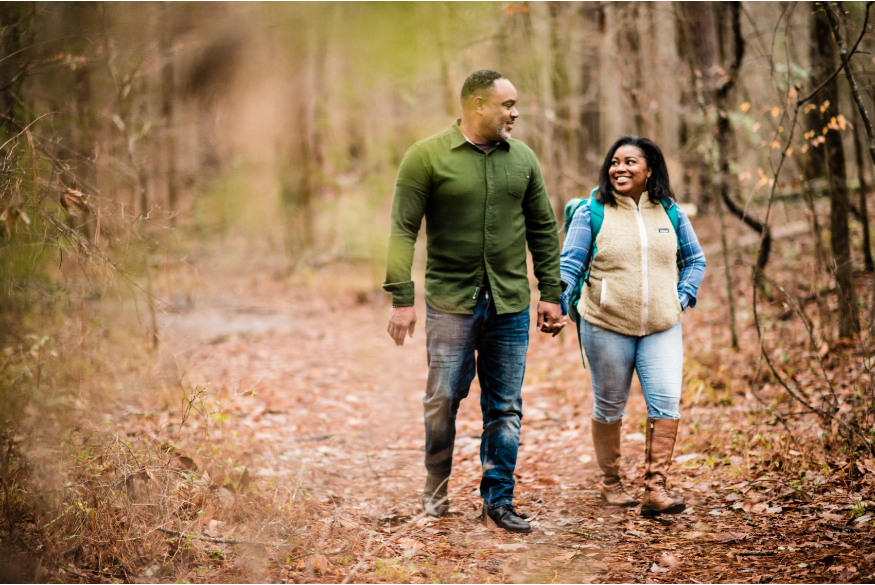 Necota and Sonya hold hands while walking on a leaf-covered trail in the forest. 