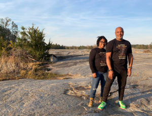 Sonya and Necota smile while standing on a large rocky plain. They are wearing matching shirts that say "camping, overlanding, rebelling, biking, diversify, hiking, rafting, kayaking, climbing."