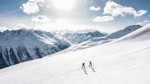 Two skiers skin up a low-angle snowfield in single file. It's sunny and mountains extend in the distance.