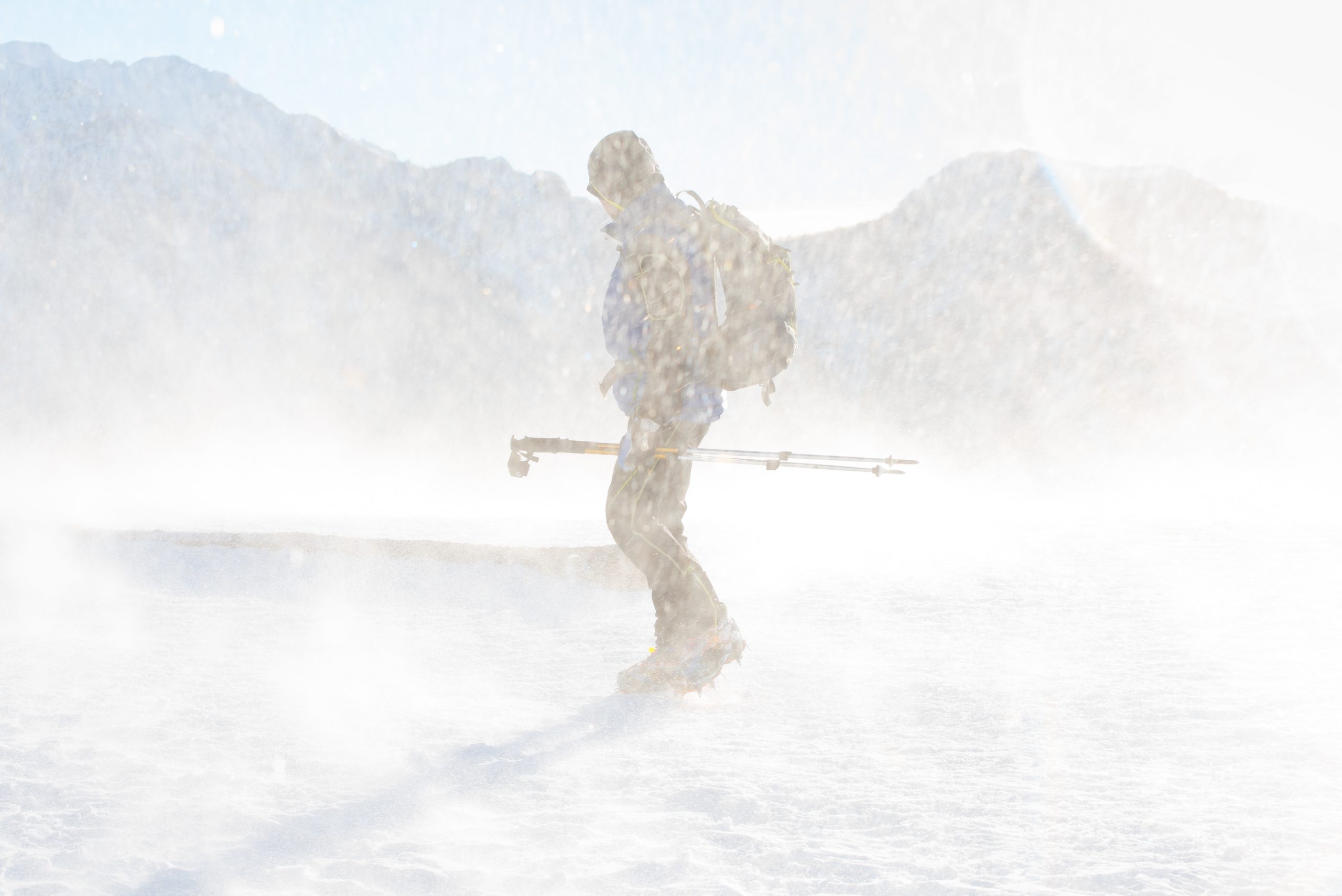 A hiker turns their face against the wind. They're carrying poles, wearing crampons, and have a backpack, hood, sunglasses, and winter coat. They're standing on a snowfield with mountains in the background.