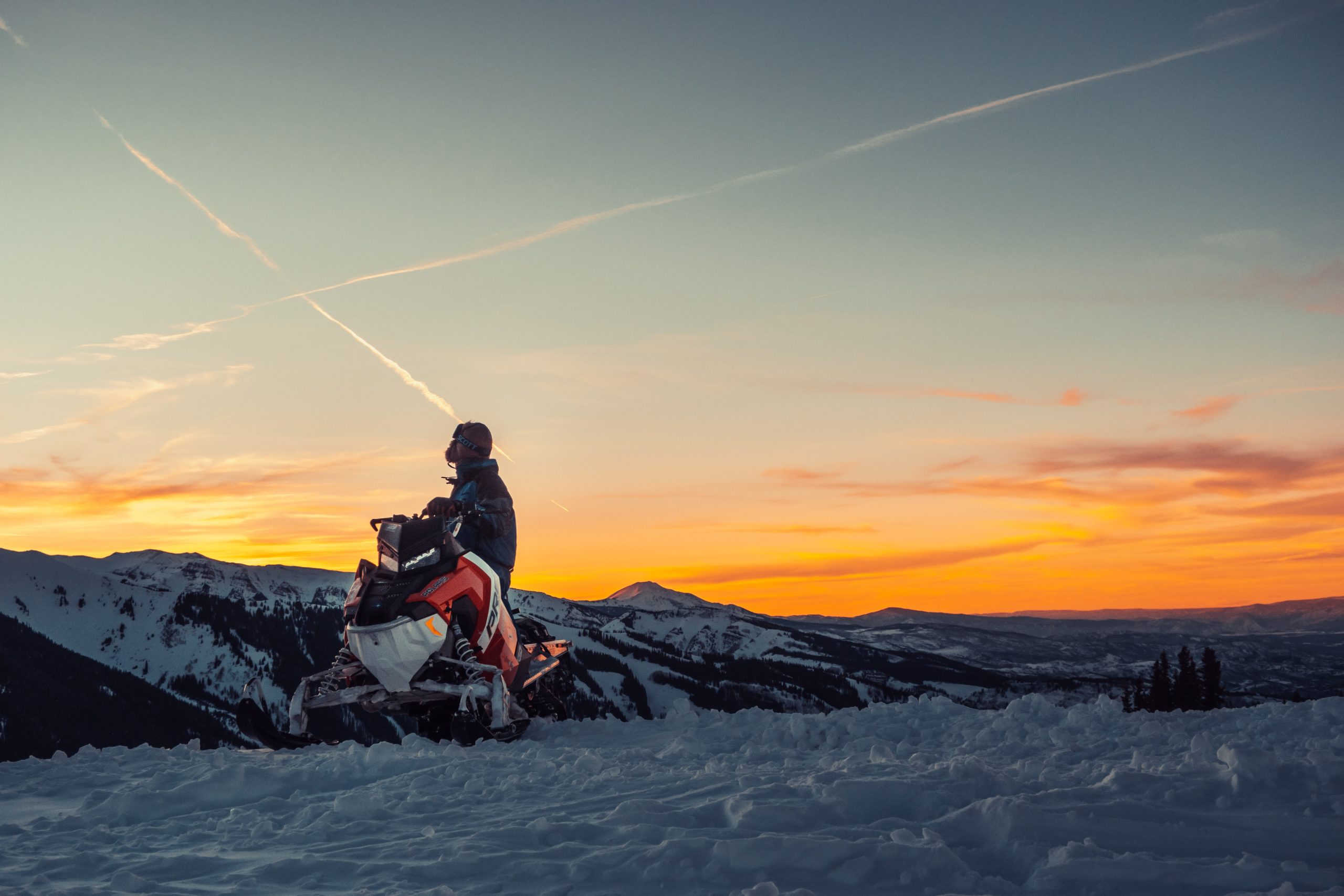 A snowmobiler looks off at the sunset over the mountains.