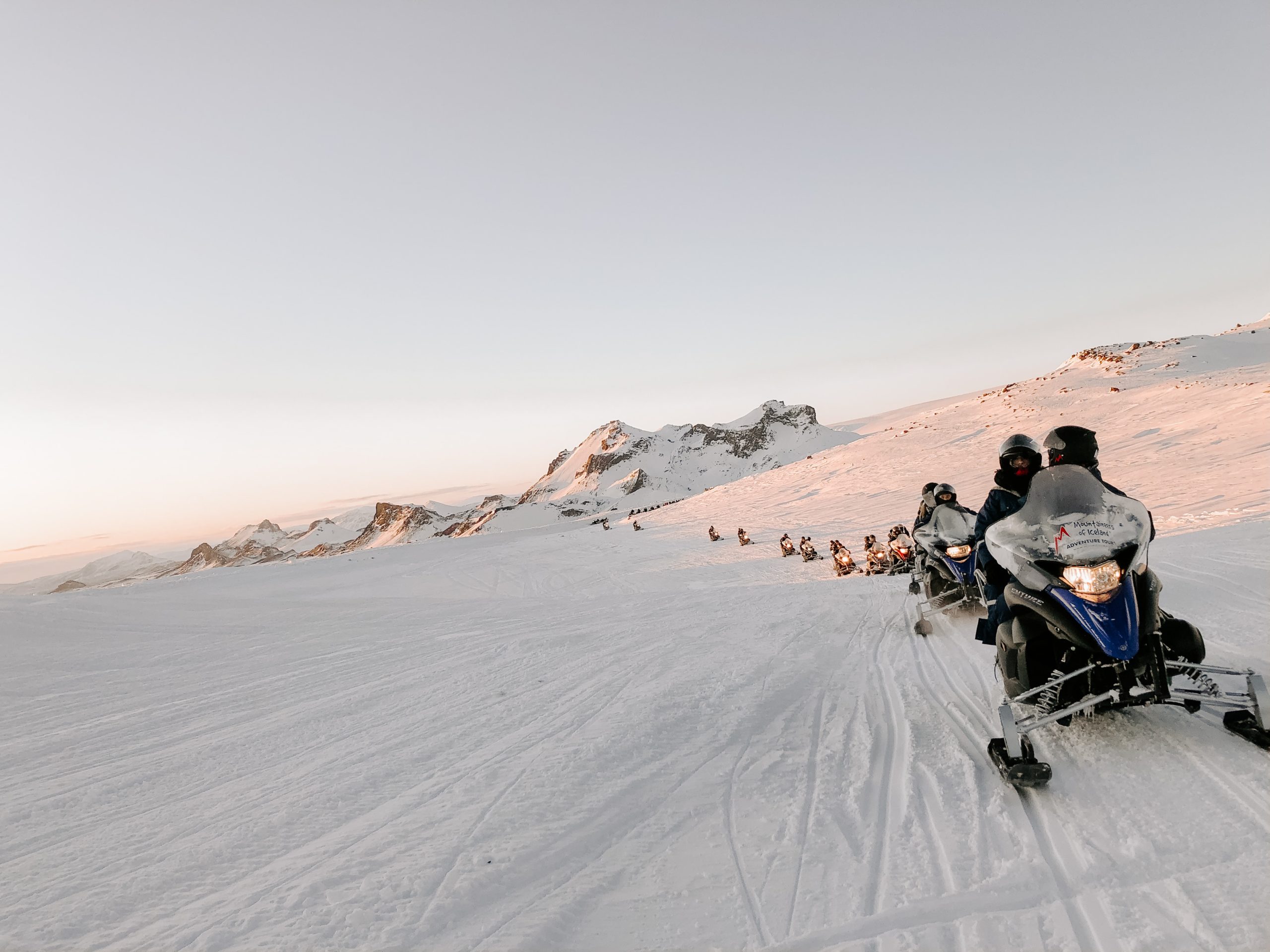 A single file line of sledders ride over a snowy plain with small mountains in the distance.