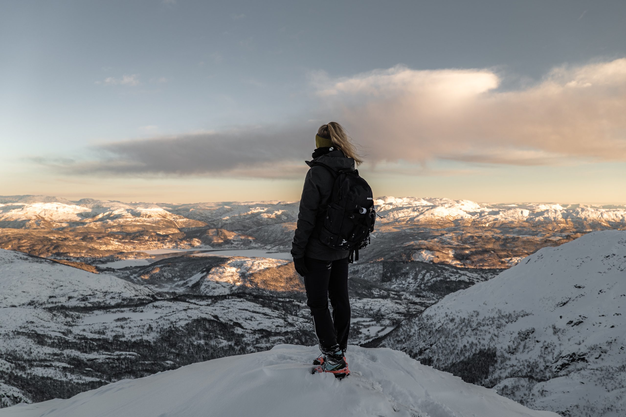 A hiker stands with their back to the camera, gazing down from the top of a mountain onto mountains, a lake, and plains covered in snow. They're wearing a headband, winter coat, and a backpack.