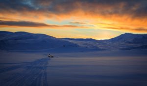 Two snowmobilers ride across a vast snow plain in the distance, in front of a glowing sunset over snowy mountains.