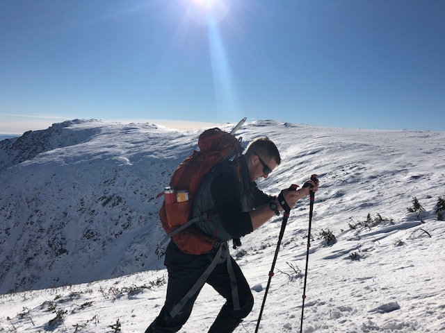 Billy hikes up near the top of Mount Washington, he's holding poles and wearing a big backpack.