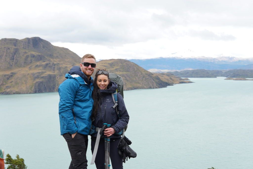 Two hikers smile while standing in front of a lake with mountains in the distance.