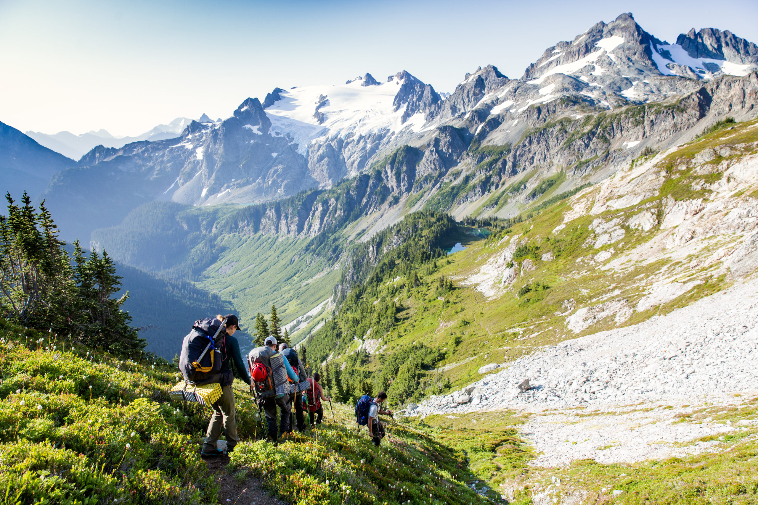A group of backpackers walk single file along a trail with mountains ahead.
