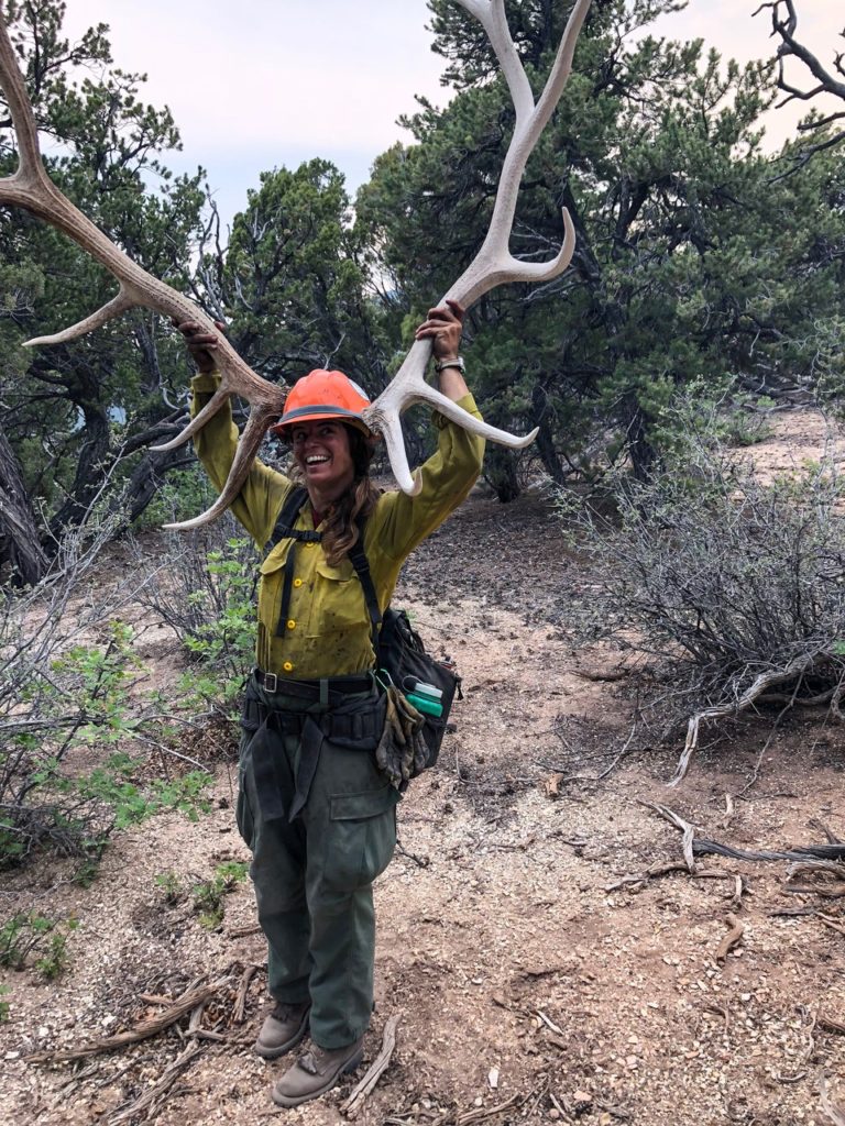 Amanda holds a giant set of antlers next to her head. She's in a hardhat and her firefighting clothes.