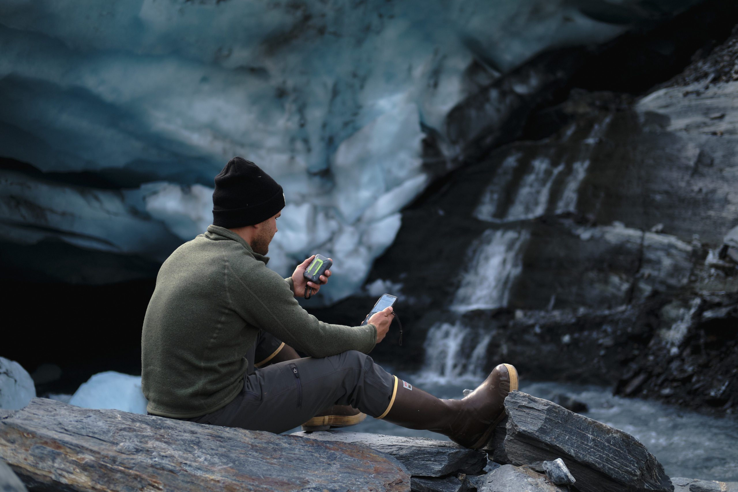 Person holds a ZOLEO in one hand and their phone in the other while sitting on some rocks.