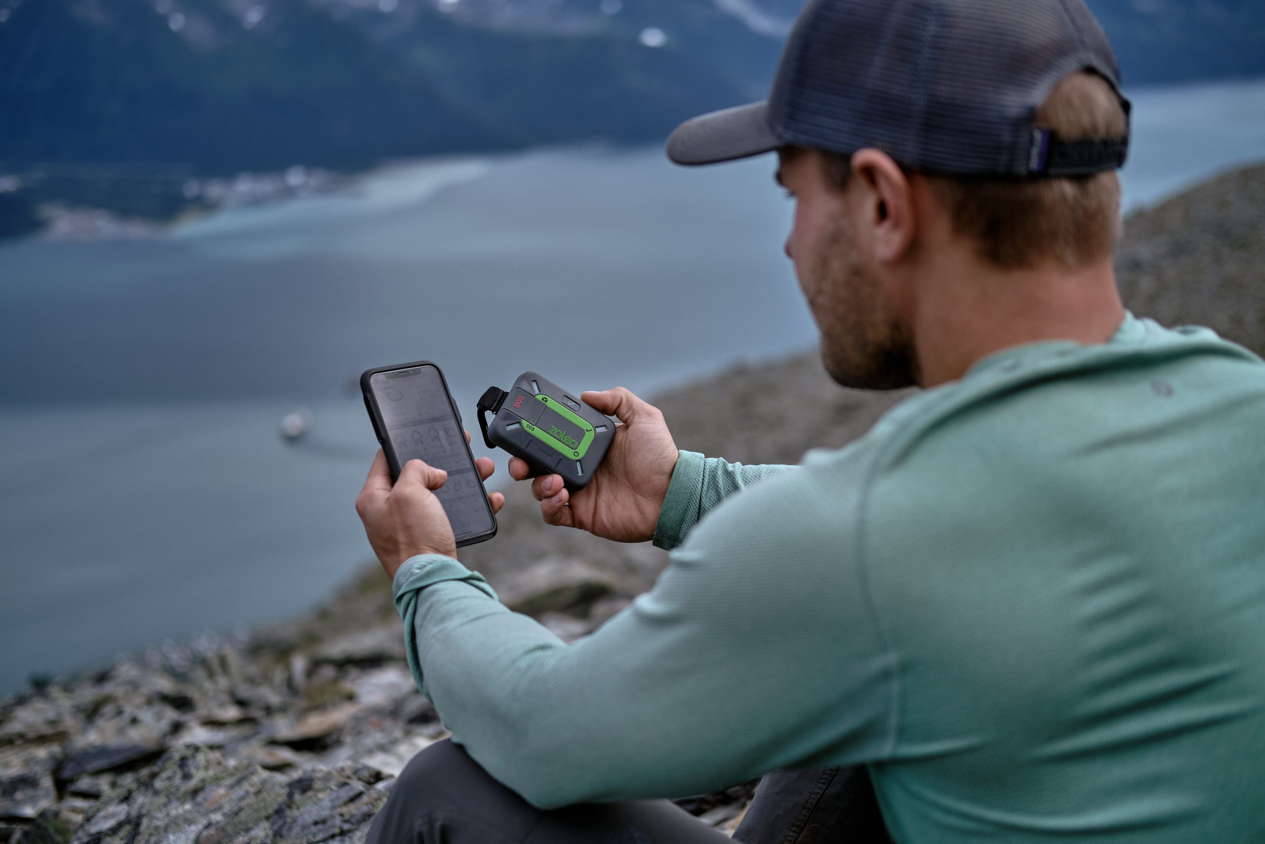 A person holds his ZOLEO unit in one hand and a phone in the other while sitting above a lake.