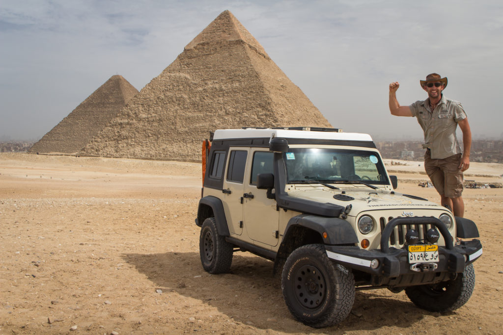 Grec stands on his Jeep tire, in front of pyramids. 