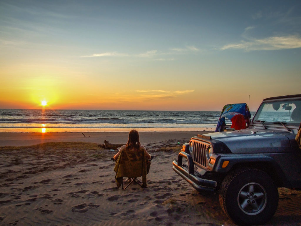 Jeep parked on a beach in front of a sunset over the ocean. 