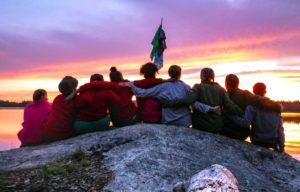 Group of kids with their arms around their backs sit on a rock looking at the sunrise.